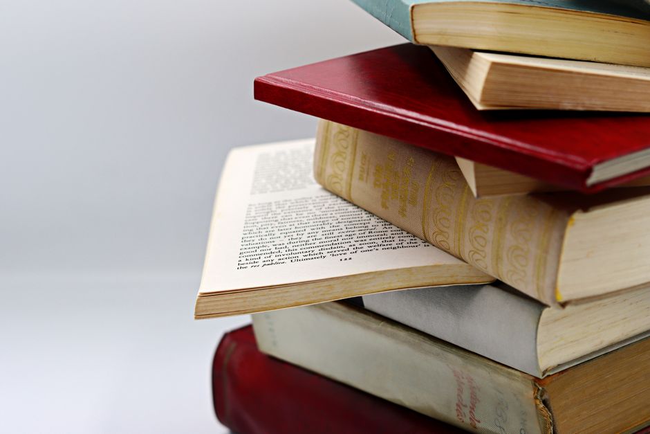 A stack of old books sits on a shelf by a sunlit window, capturing a sense of quiet study.